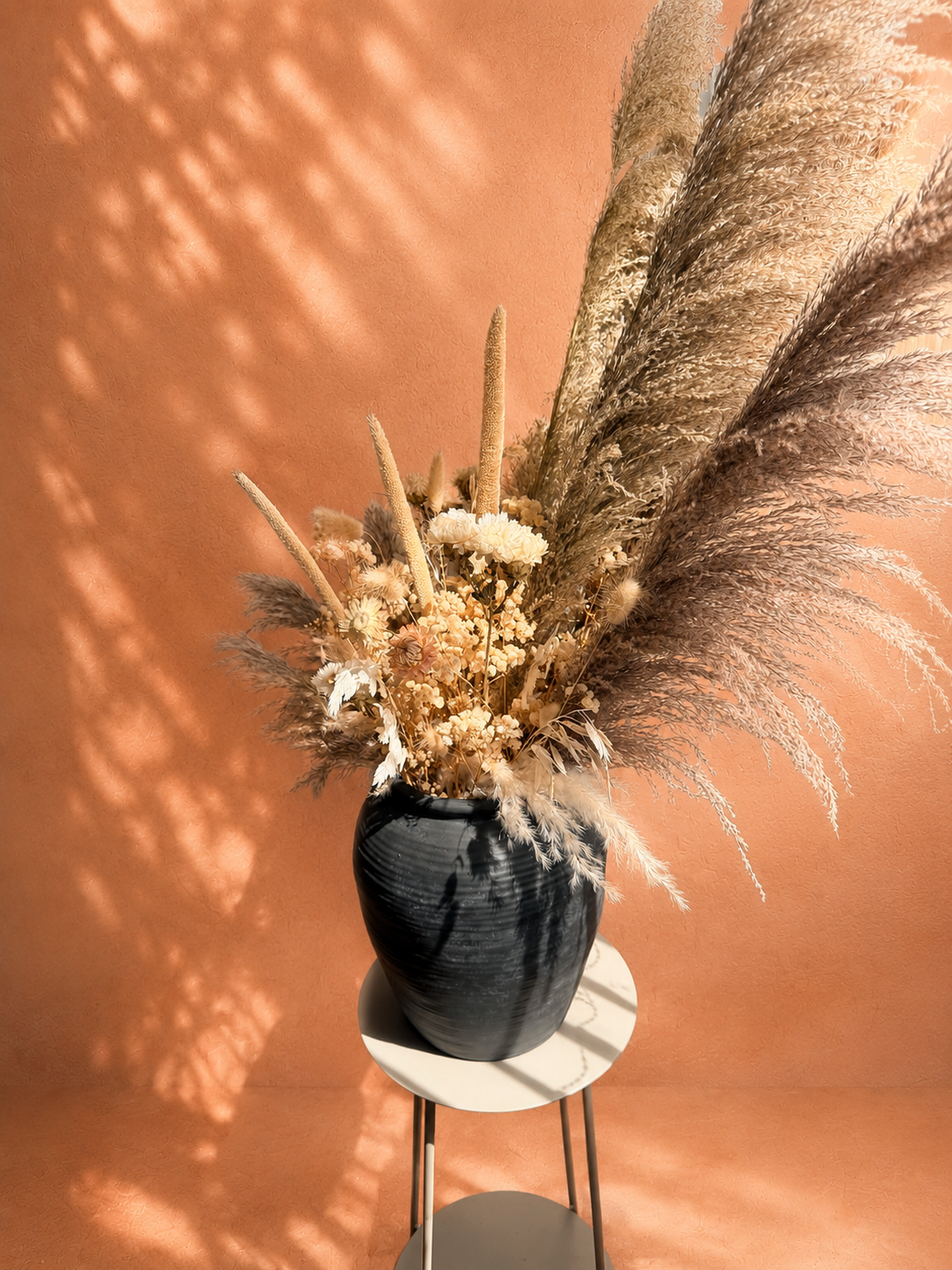 Pampas Mixbunt 'TRE KRONOR' med pampas och torkade blommor i beige, gråbruna och vita toner, arrangerad i en svart vas mot en terrakottafärgad vägg.
