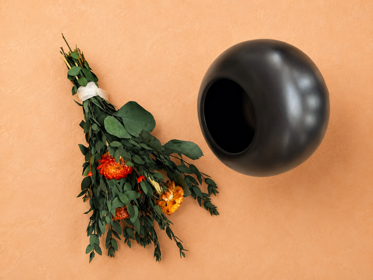 Svart rund vas med bred öppning bredvid en bunden bukett av gröna blad och orange torkade blommor på aprikosfärgad bakgrund.
