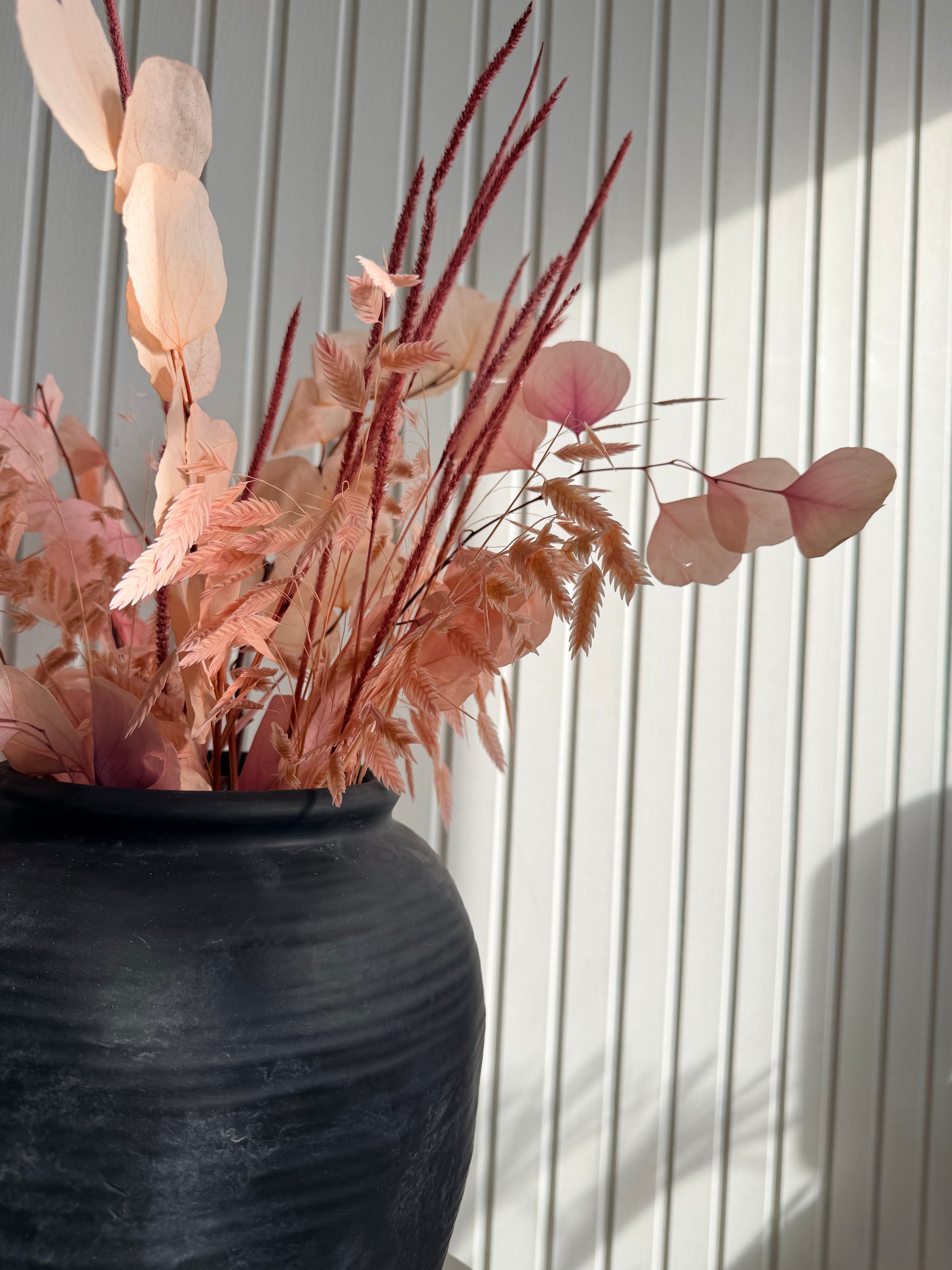 Torkad och bevarad bukett i mjuka rosa toner i svart keramikvas, soligt fönsterljus mot vit panelvägg, modern skandinavisk inredning, torkade blommor och pampas från Torrdecor