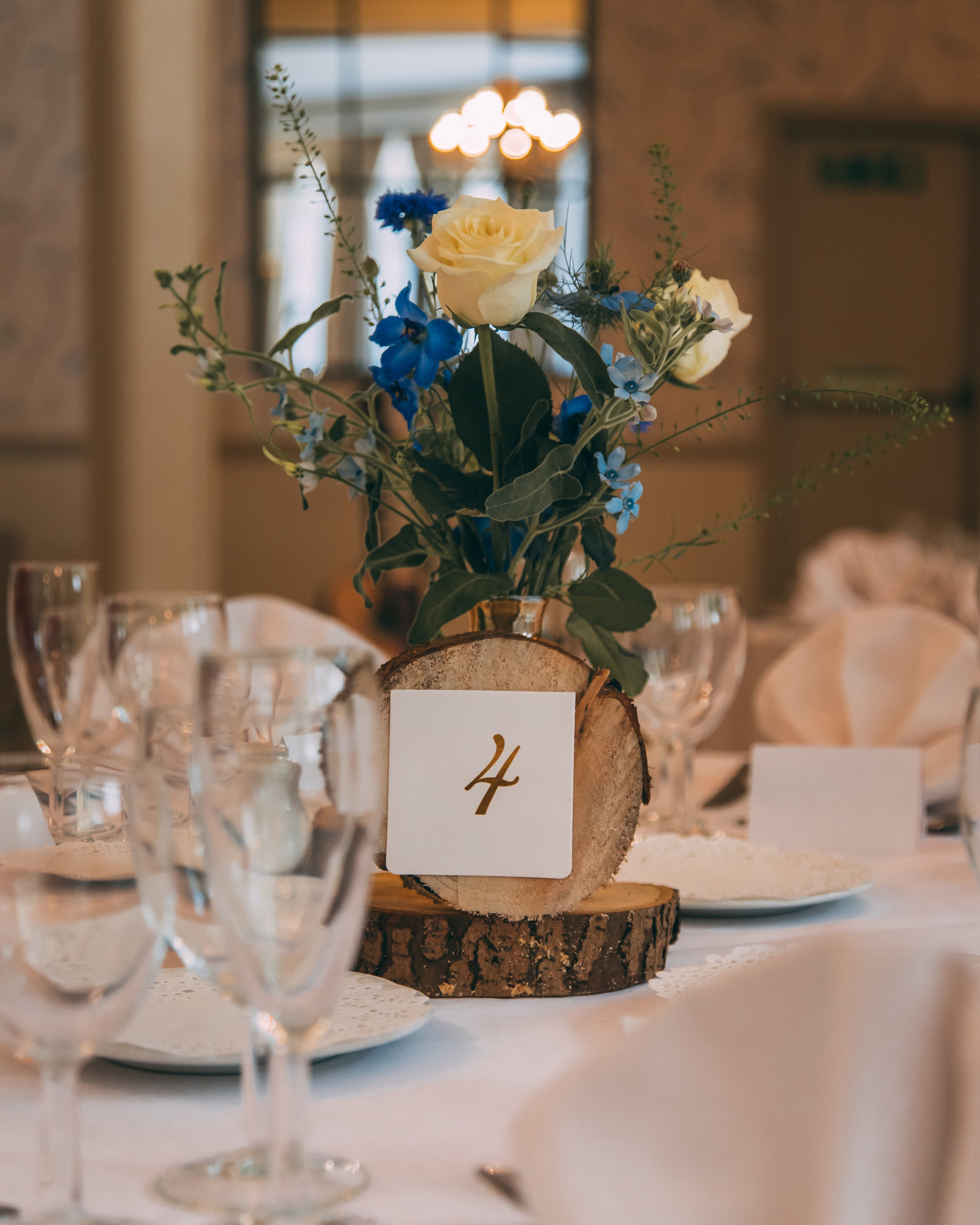 Elegant bordsdekoration med snittblommor på dukat festbord med vit ros och blå blommor i glasvas samt bordsnummer på träskiva vid bröllop eller middag